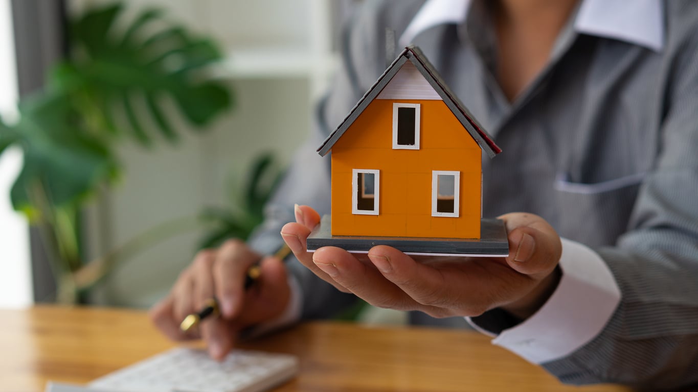 concept of expensive home in current real estate market with high pricing; real estate agent holding out model home in hand concept of expensive home in current real estate market with high pricing; real estate agent holding out model home in hand