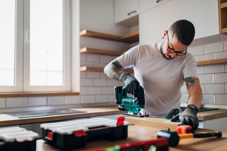 The bearded carpenter using electric jigsaw for cutting a wooden plank in his new kitchen. Home Remodel Purchase Mortgage