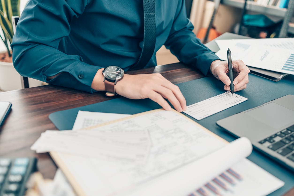 Business man hand writing and signing white blank bank check book and dollar bill, coin, laptop and graph chart on the desk at office. Payment by check, paycheck, payroll concept.